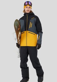 Kurtka snowboardowa w czerni i żółci z szarymi akcentami, z zamykanymi kieszeniami, w komplecie z dopasowanymi czarnymi spodniami i deską snowboardową.