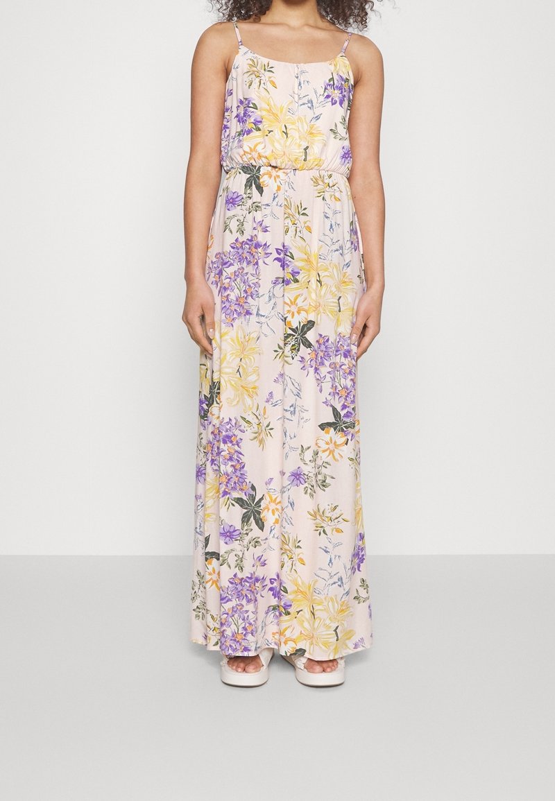 Femme portant une robe sans manches longue jusqu’au sol avec des motifs floraux jaunes, violets et verts sur un fond clair et des sandales blanches.