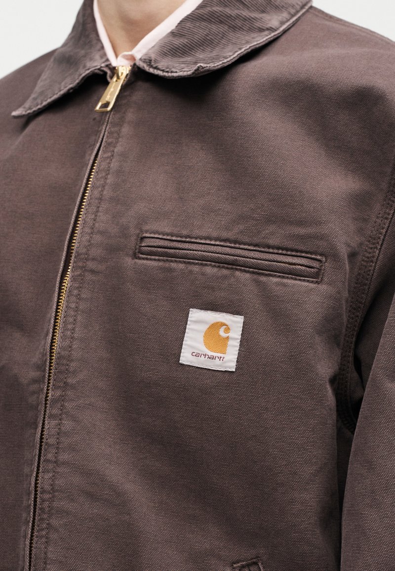 Primer plano de chaqueta Carhartt marrón con cremallera visible, cuello, bolsillo en el pecho y parche con logo blanco en el lado izquierdo.
