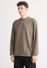 Chemise à manches longues en coton vert olive avec un col rond et un petit logo sur la poitrine, présentant des manches à poignet et une coupe décontractée.