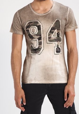 Camiseta de manga corta color marrón claro con un acabado desgastado que presenta una gran impresión de camuflaje "94" en la parte delantera. Textura de tela suave.