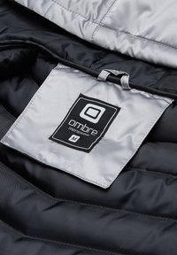Svart och grått pufferjacka interiör som visar en skötselrådsetikett med en "ombre menswear"-logotyp, broderad på ett slätt metallgrått tyg.