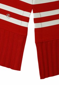 Close-up van rood en wit gestreepte gebreide mouwen met geribbelde manchetten en een kleine ronde zilveren logo-pin aan één kant.