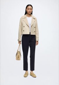 Femme portant un trench-coat beige court, un top blanc, un pantalon noir, des mocassins beiges et tenant un sac à main beige sur fond blanc.