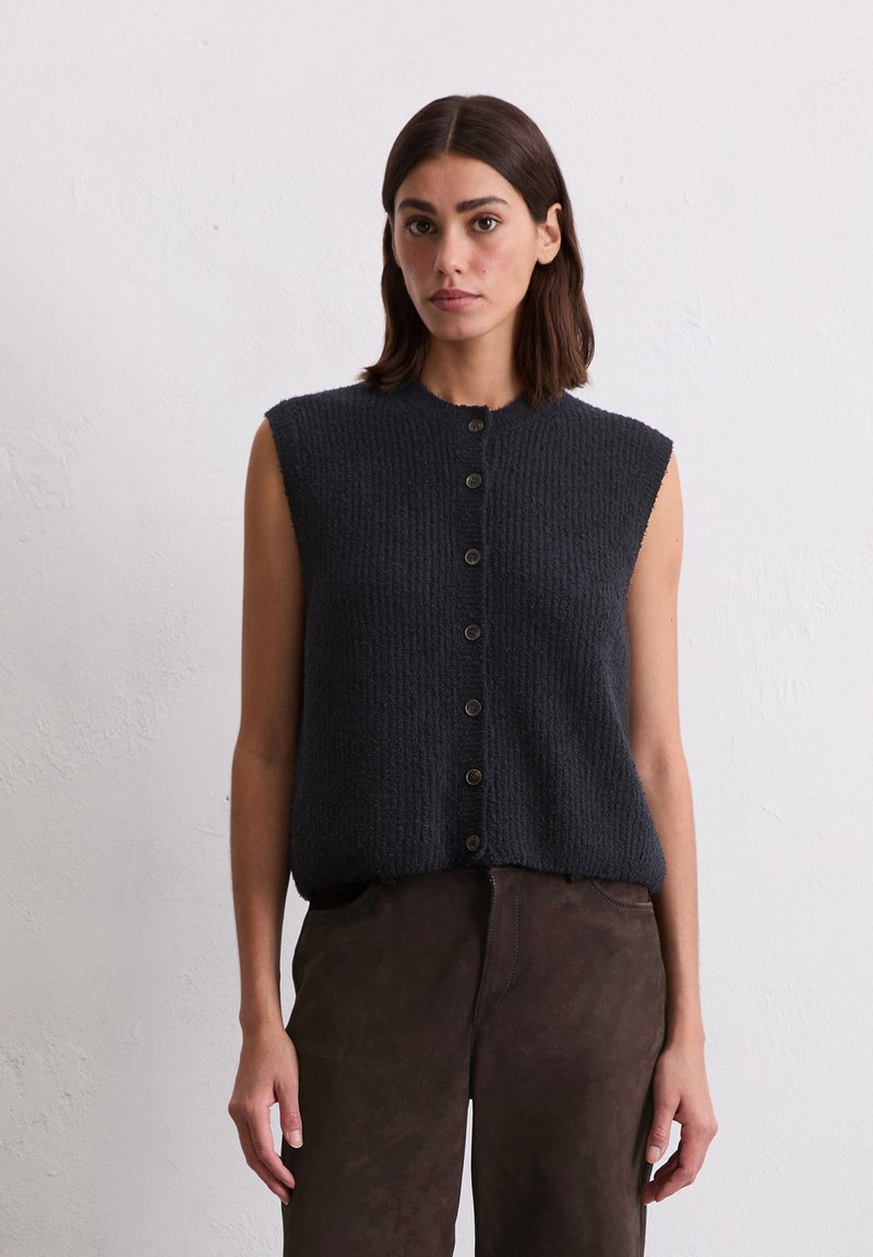 Top sans manches tricoté gris foncé avec une finition texturée, encolure ronde et boutons à l'avant. Associé à un pantalon ajusté marron.