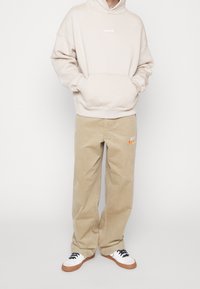 Personne portant un sweat à capuche beige avec poche avant, un pantalon ample kaki avec le logo Nike, et des baskets blanches avec semelles en gomme et lacets noirs.
