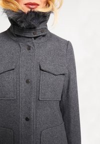 Cappotto di lana grigio con un colletto alto e bordo in pelliccia, due tasche sul petto, quattro bottoni sul davanti e una finitura texturizzata.