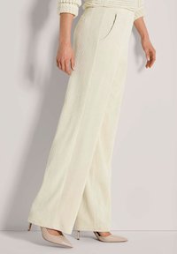 Vrouw in een crèmekleurige corduroy broek met wijde pijpen en beige puntige hoge hakken, staand tegen een lichte achtergrond.