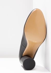 Chaussure à talon haut en cuir noir avec une tige texturée, un bout arrondi et un talon cylindrique avec semelle en caoutchouc. L'intérieur affiche les marquages de taille.