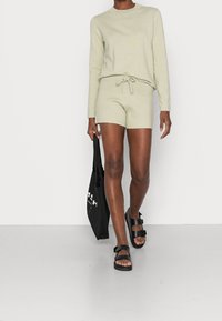 Jasnozielony zestaw dresowy składający się z długiego rękawa bluzki i szortów. Posiada okrągły dekolt, regulowany pasek w talii oraz luźny krój. Noszony z czarnymi sandałami.