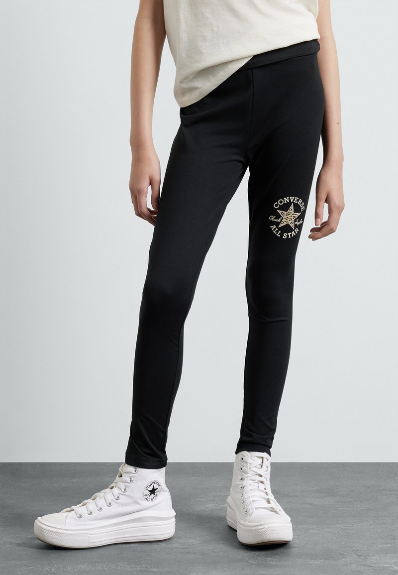 Leggings neri con un design aderente, caratterizzati da un logo dorato "Converse All Star" sulla gamba sinistra. Abbinati a sneakers alte bianche.