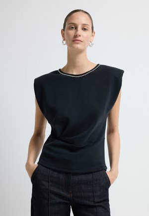 Top noir sans manches avec un col rond, orné d'une accent en perles. Tissu doux, forme sculpturale et subtil détail de drapé.