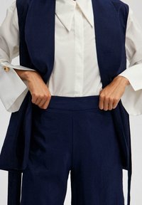 Mörkblå, strukturerade byxor som matchas med en vit skjorta med stor krage; modellen justerar midjebandet med händerna.