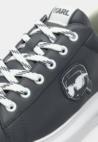 Σκούρα γκρι δερμάτινο αθλητικό παπούτσι με λευκά και μαύρα κορδόνια με μοτίβο, διαθέτει λευκό λογότυπο και λαστιχένια σόλα. Λεπτομέρεια υφής στην επιφάνεια.