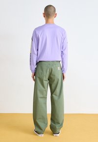 Osoba z ogolona głową, ubrana w lawendową bluzkę z długim rękawem z tekstem na rękawie, zielone spodnie w szerokim kroju i czarno-białe sneakersy, stoi.