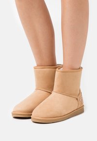 Paire de bottes mi-mollet en daim beige avec semelles plates et doublure douce, portées sur des jambes nues, posées sur un fond blanc uni.