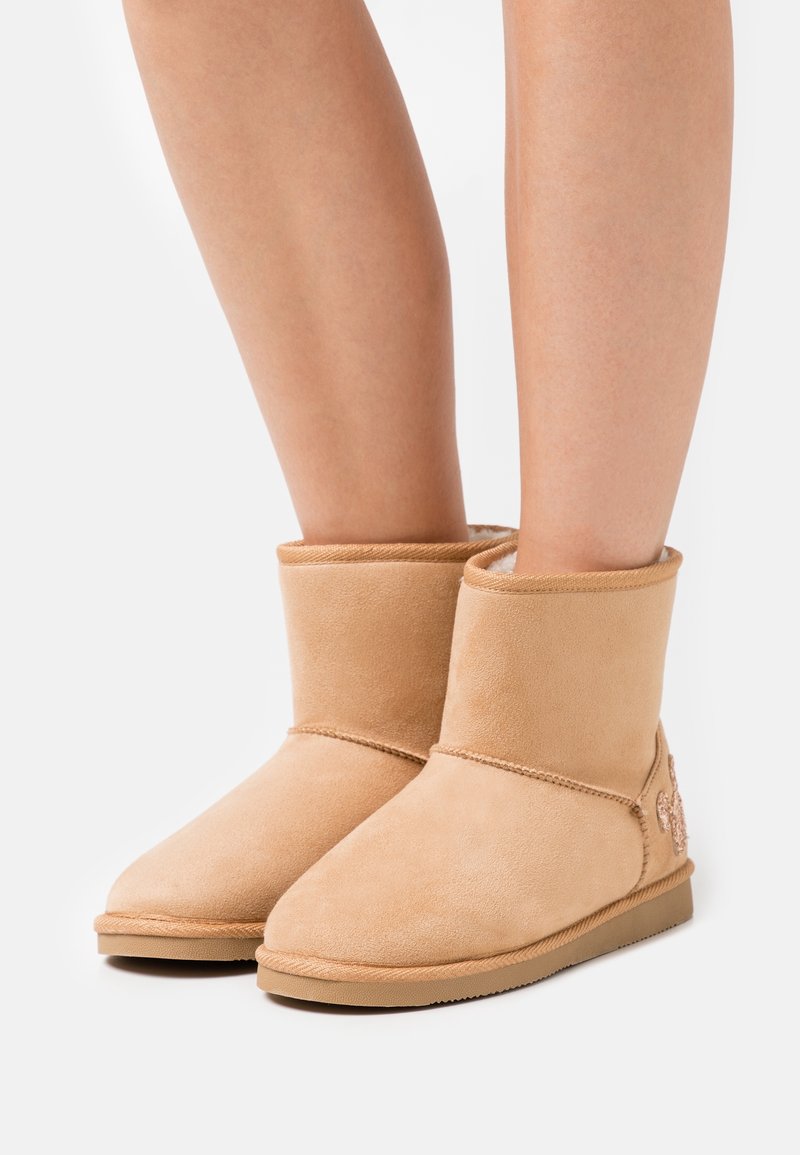 Paire de bottes mi-mollet en daim beige avec semelles plates et doublure douce, portées sur des jambes nues, posées sur un fond blanc uni.