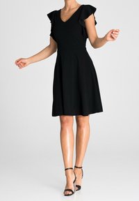 Vestido negro con escote en V, mangas cortas con volantes, corsé ajustado y falda acampanada. Lleva unos tacones negros de tiras. Textura de tela suave.