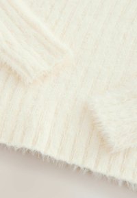 Tissu tricoté de couleur crème clair avec une texture douce et duveteuse. Présente un motif côtelé, avec des bords légèrement effilochés au niveau des poignets.