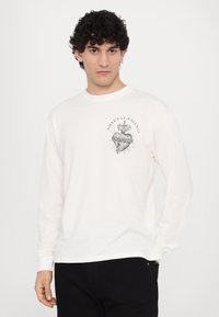 Tricou alb din bumbac cu mânecă lungă, având un design grafica negru în formă de inimă cu raze și o mână în zona pieptului. Textură netedă.