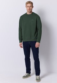 Grüner Pullover mit Rundhalsausschnitt, Rippbündchen und Saum. Getragen mit dunkelblauen Jeans und hellgrünen Turnschuhen, die ein Logo auf der Brust haben.
