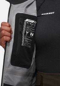 Svart Mammut-jackas inre med en zipficka som visar säkerhetsinstruktioner, metalliska detaljer och ett grått tygfoder.