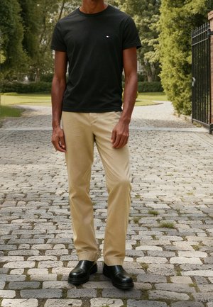 Man staat op een geplaveid pad, gekleed in een zwart t-shirt, beige broek en zwarte leren schoenen, met groene bomen op de achtergrond.