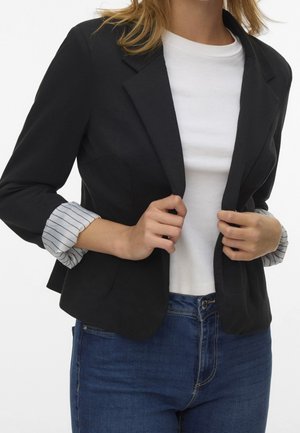 Woman wearing a black blazer with rolled striped cuffs over a white t-shirt and blue jeans, holding the blazer’s front edges.