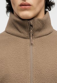 Άτομο ντυμένο με καφέ φλις ζακέτα με φερμουάρ κλειστό μέχρι ψηλά, με γειαλόνι που καλύπτει τον λαιμό, εμφανίζοντας μόνο το κάτω μέρος του προσώπου.