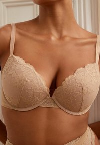 Beige Spitzen-BH mit floralem Muster, gepolsterten Cups, verstellbaren Trägern und glatter Textur. Verfügt über einen tiefen Ausschnitt und gewellte Kanten.