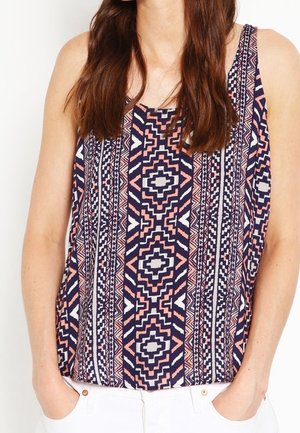 Mouwloos topje met marineblauwe, roze en witte geometrische tribale patronen, gedragen met witte broek, handen in de zakken.