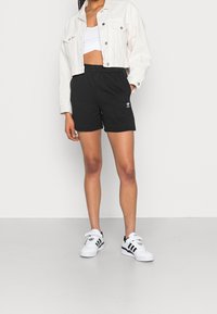 Svarta idrottsshorts med elastisk midja och vitt logotyp, tillsammans med en cropped vit denimjacka och vita sneakers med svarta ränder.