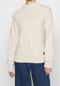 Pull beige avec un col montant côtelé et un motif texturé. Caractérisé par des poignets et un ourlet en côtes épaisses, associé à un jean en denim bleu.