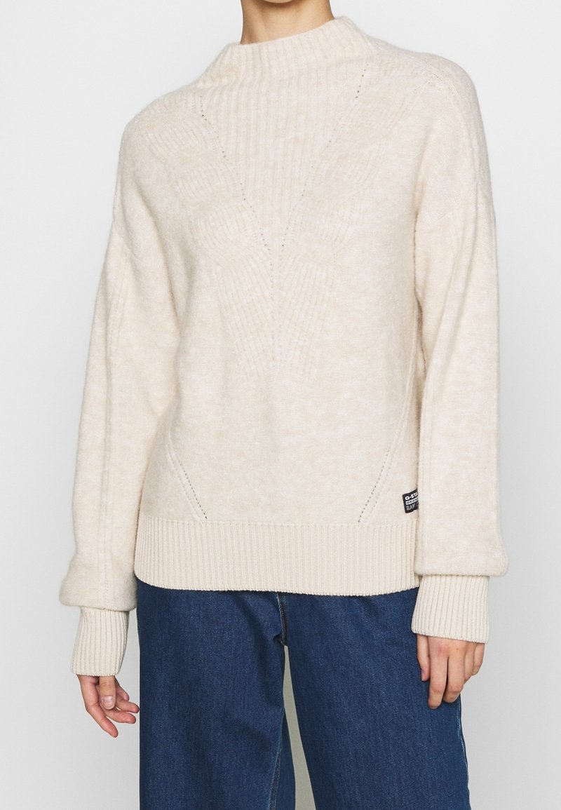 Pull beige avec un col montant côtelé et un motif texturé. Caractérisé par des poignets et un ourlet en côtes épaisses, associé à un jean en denim bleu.