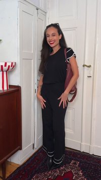 Femme aux longs cheveux foncés, tenue noire avec bordure blanche, rouge à lèvres rouge et sac rouge foncé, souriante debout dans une pièce avec des portes blanches et un tapis à motifs.