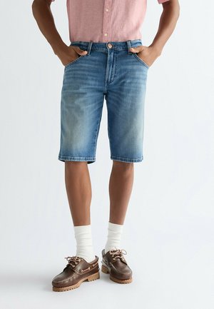 Blå denimshorts med whiskering, design med fem lommer og en afslappet pasform. Bruges med brune sko og hvide sokker, mod en ensfarvet baggrund.