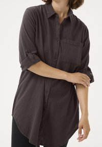 Chemise button-up gris foncé à manches longues retroussées aux coudes, avec une poche poitrine unique et un ourlet arrondi. Texture douce, tissu léger.