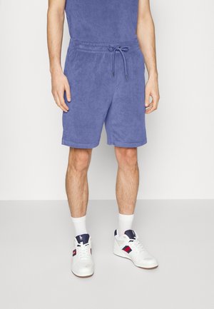 Mężczyzna w niebieskich szortach z froty, białych skarpetkach i białych trampkach z granatowymi i czerwonymi akcentami stoi na jednolitym tle.