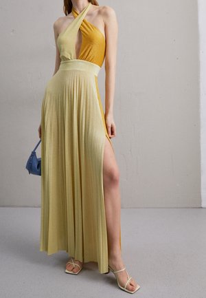 Mujer con un vestido halter amarillo y dorado con falda plisada y abertura alta en el muslo, lleva un bolso azul y sandalias de tiras.