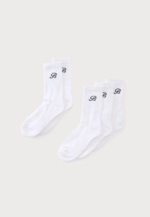 Deux paires et trois paires de chaussettes blanches montantes avec un logo "B" noir en cursive sur les bords côtelés, disposées sur un fond clair.