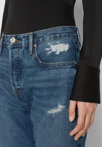 Jean en denim bleu avec une taille haute, des zones usées et des surpiqûres contrastantes. Une fermeture à bouton et braguette se trouve à la taille.