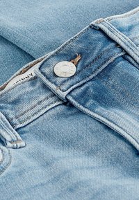 Jeans de mezclilla azul claro con cierre de botón plateado, costuras en contraste y una textura de tela visible con ligeras marcas de desgaste.