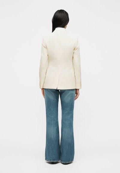 Γυναίκα με μακριά σκουρόχρωμα μαλλιά, φοράει εκρού σακάκι και μπλε τζιν παντελόνι φαρδιάς γραμμής, στέκεται με την πλάτη γυρισμένη σε ένα απλό λευκό φόντο.