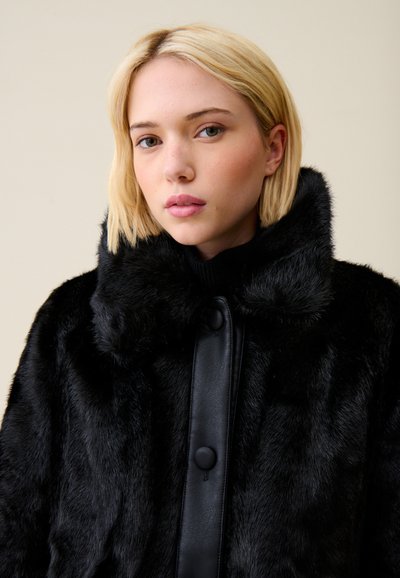 Giovane donna con capelli biondi corti che indossa un cappotto in pelliccia sintetica nera con dettagli in pelle, su uno sfondo uniforme chiaro.