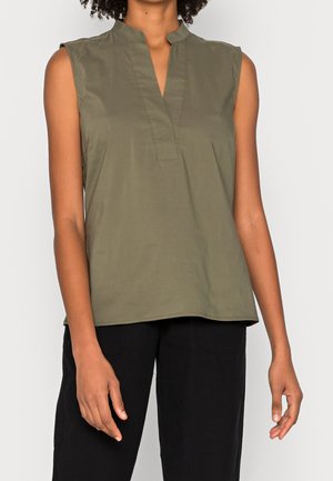 Femme portant un haut sans manches olive avec un col en V et un pantalon noir, se tenant devant un fond uni clair.