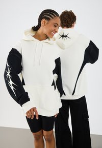 Witte en zwarte hoodie met een kangoeroezak; heeft grote zwarte accenten en een abstract wit patroon op een mouw.