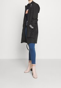 Giacca nera con cappuccio e coulisse, con scritta bianca sulla manica, abbinata a jeans skinny blu e stivaletti beige.