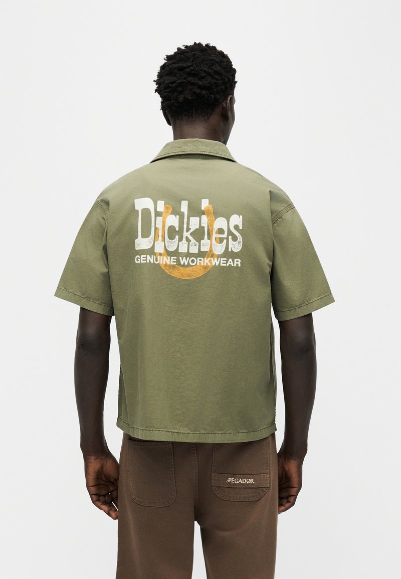 Hombre vistiendo una camisa de manga corta verde oliva con el logo "Dickies Genuine Workwear" en la espalda y pantalones marrones con parche en el bolsillo trasero.