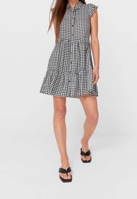 Frau trägt ein ärmelloses, schwarz-weißes Karo-Kleid mit Knopfverschluss, Rüschensäumen und schwarzen Sandalen mit quadratischer Zehenpartie.