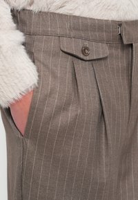 Hnedé nohavice s tenkými prúžkami a záhybmi vpredu, vybavené zapínaním na gombík a bočnými vreckami. Materiál má jemný, textúrovaný povrch.